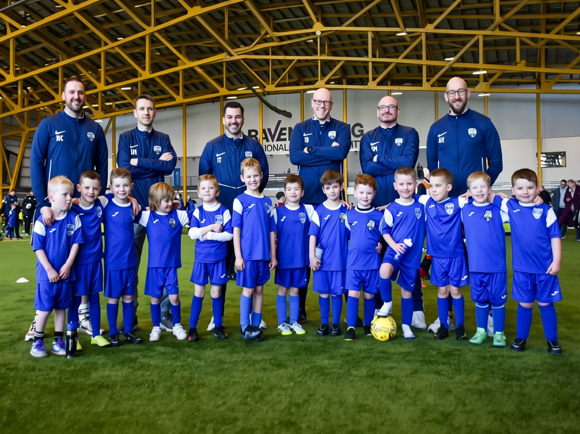 A group of young boys and their coaches posing for a team photo inside an indoor sports facility. The boys are wearing blue soccer uniforms, and the coaches are standing behind them in matching blue tracksuits.