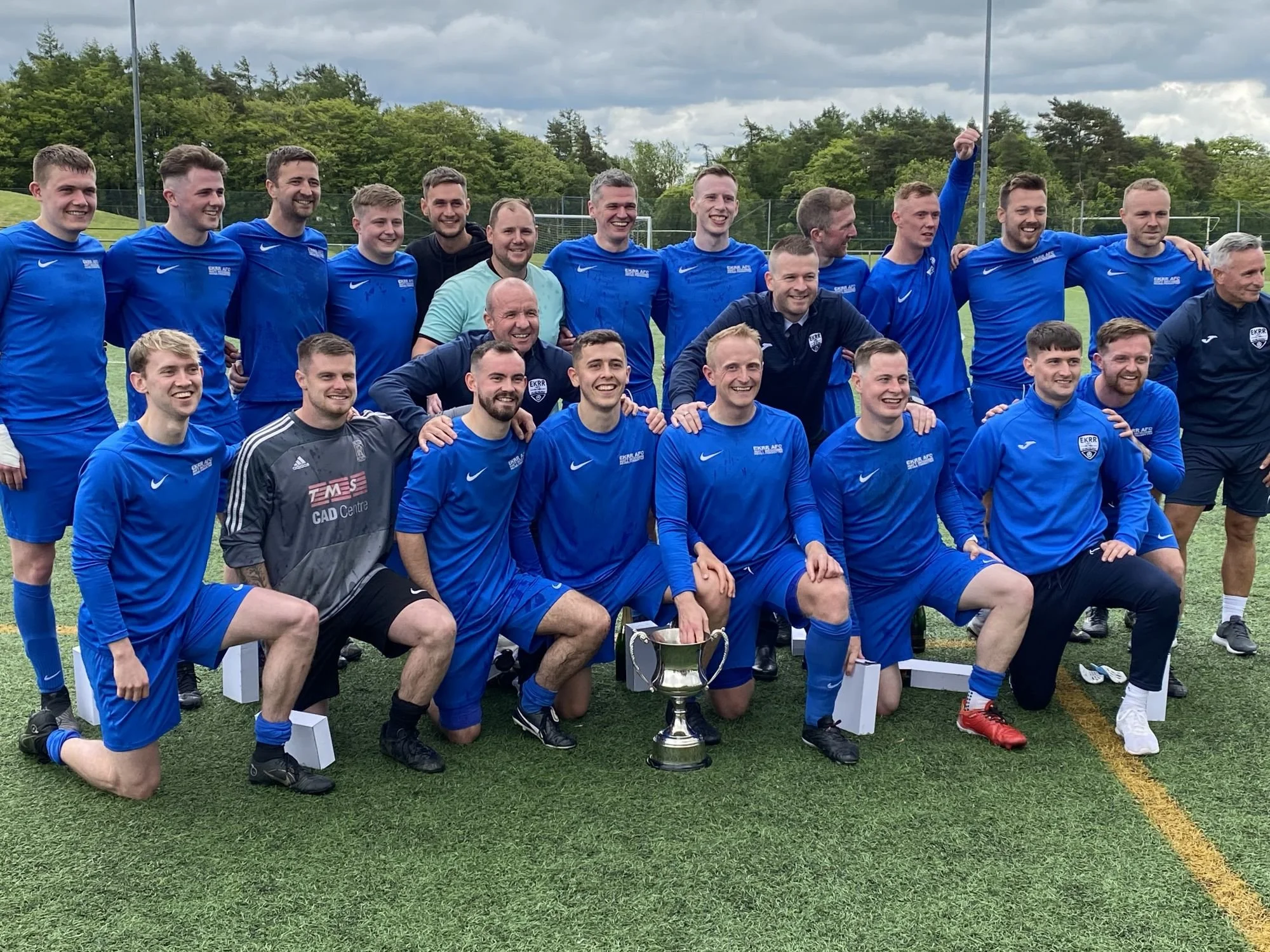 A group of young men in blue soccer uniforms posing with a trophy on a soccer field during daytime. They are smiling, some kneeling, and others standing behind them, with trees and cloudy sky in the background.