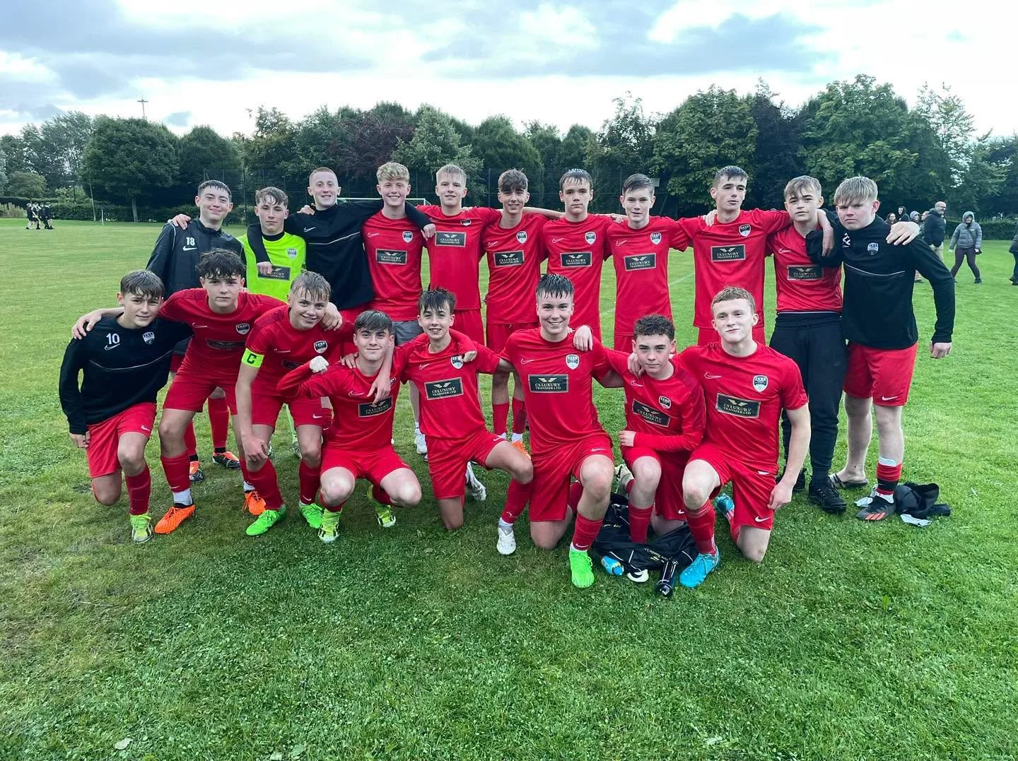 Young male soccer players in red uniforms and black jackets posing on a grassy field after a game, with trees and cloudy sky in the background.