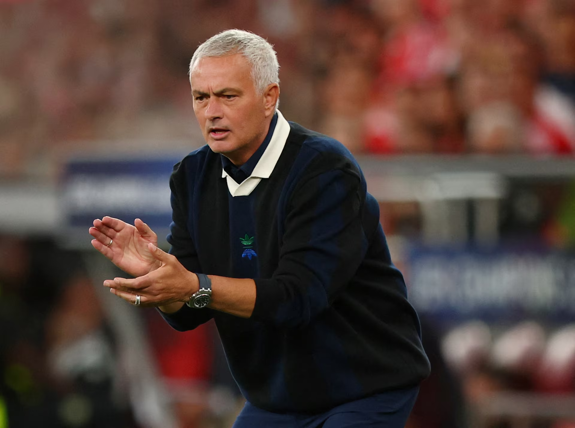 A man with short gray hair, wearing a dark jacket, clapping his hands, on a sports field with blurred spectators in the background.