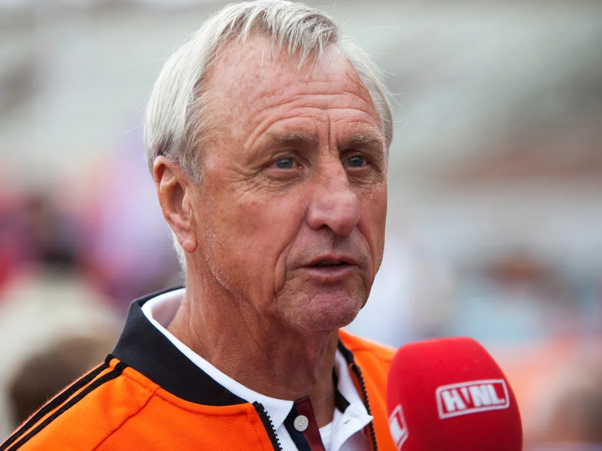 An older man with short, light-colored hair being interviewed outdoors, holding a red microphone with the logo 'HNL' on it. He is wearing an orange jacket with black stripes on the shoulders.