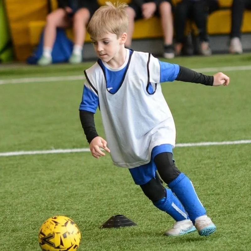 Young boy playing soccer on indoor field, dribbling yellow ball, wearing blue jersey, black knee pads, and blue socks.