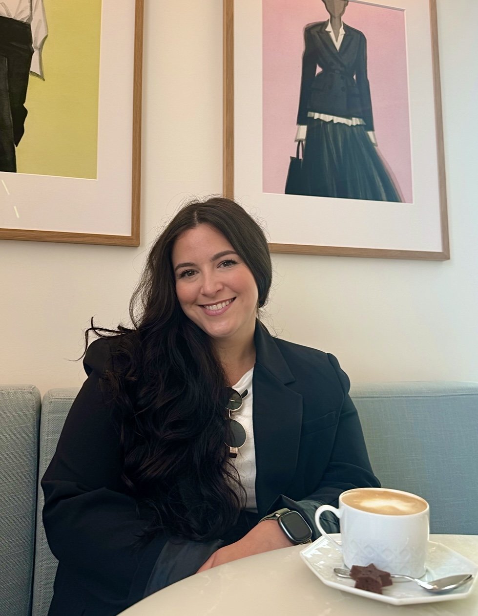 Smiling woman with long dark hair sitting at a table with a cup of coffee and dessert, in a cafe with framed fashion artwork on the wall.