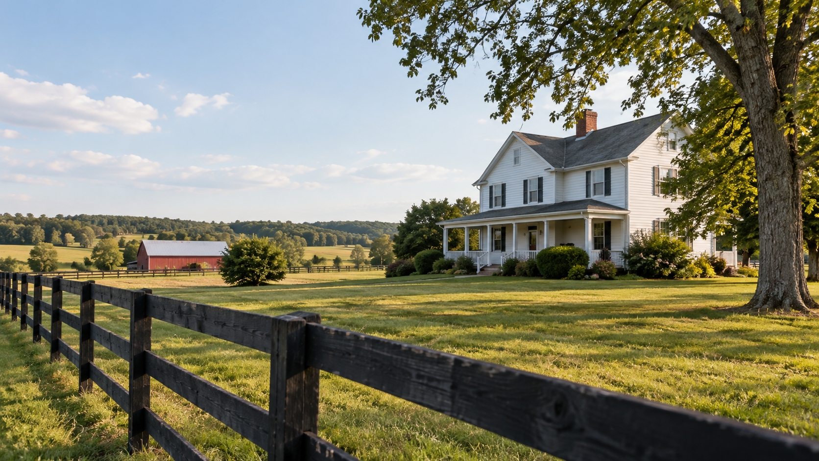 A white two-story farmhouse with a front porch, surrounded by a manicured lawn and bushes, and a large tree in the foreground. A black wooden fence runs across the bottom of the image, with a red barn and rolling hills in the background under a partly cloudy sky.