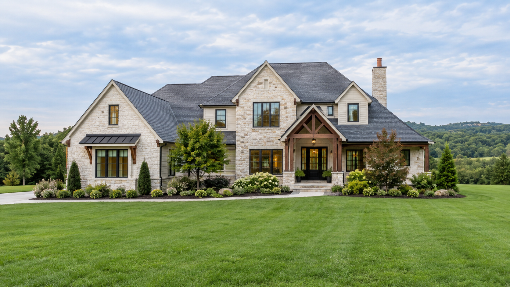 Front view of a large modern house with stone and siding exterior, surrounded by a well-maintained lawn and landscaped garden, with trees and hills in the background.