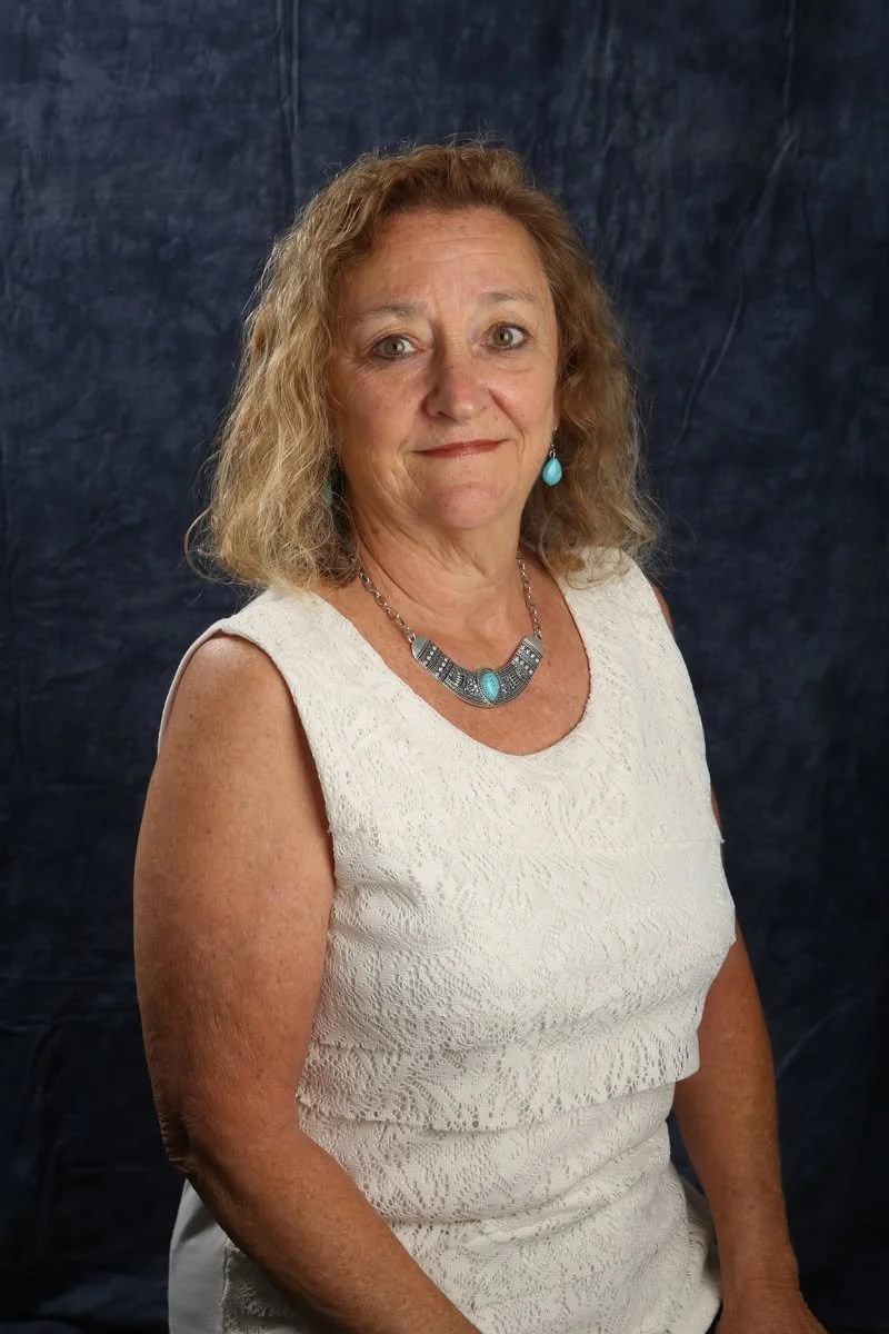 A woman with blonde, curly hair wearing a white sleeveless lace dress, turquoise jewelry, and sitting in front of a dark blue background.