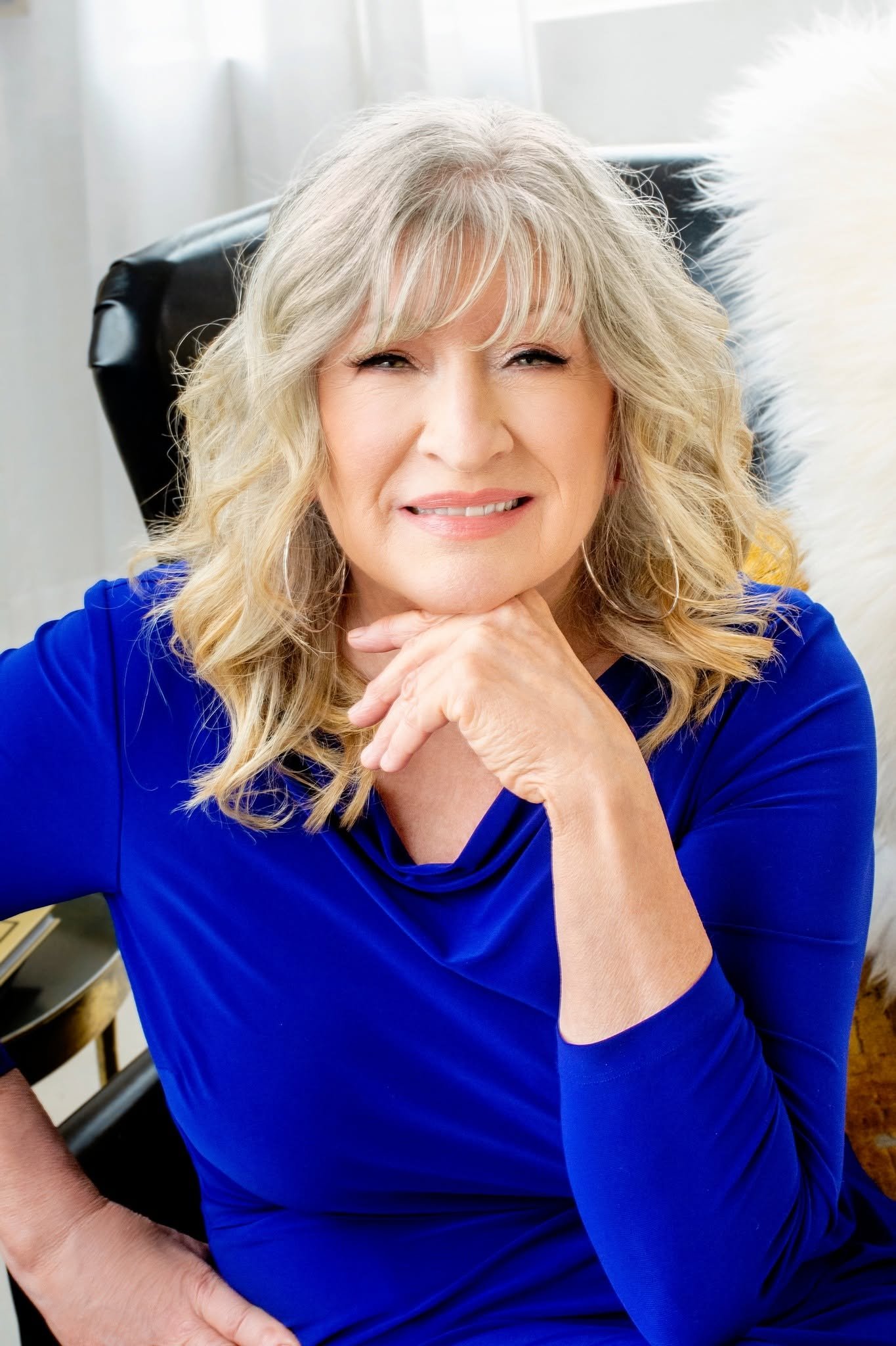 A smiling mature woman with blonde hair, wearing a royal blue dress, sitting on a chair with her chin resting on her hand and looking at the camera.