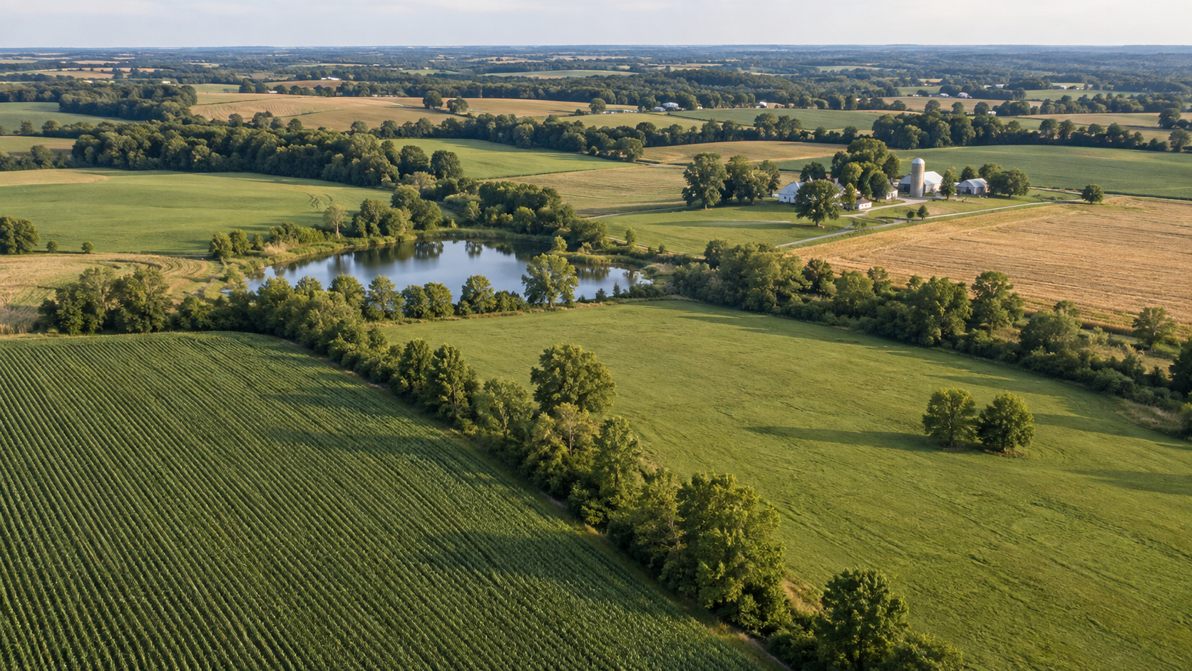 Aerial view of farmland with fields, trees, a pond, and farm buildings in the distance.