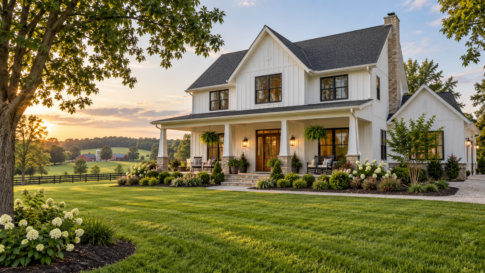 A modern white two-story house with a front porch, surrounded by a well-maintained garden and lush green lawn, with trees and a rural landscape in the background during sunset.