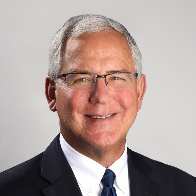 A professional headshot of an older man with gray hair, glasses, wearing a dark suit, white shirt, and blue tie.
