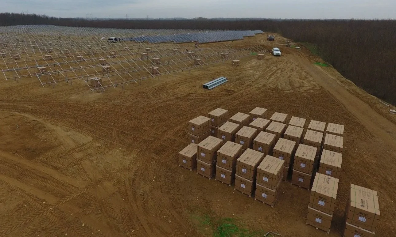 A large construction site with metal framework for solar panels, stacks of boxes, construction vehicles, and cleared dirt paths surrounded by trees.