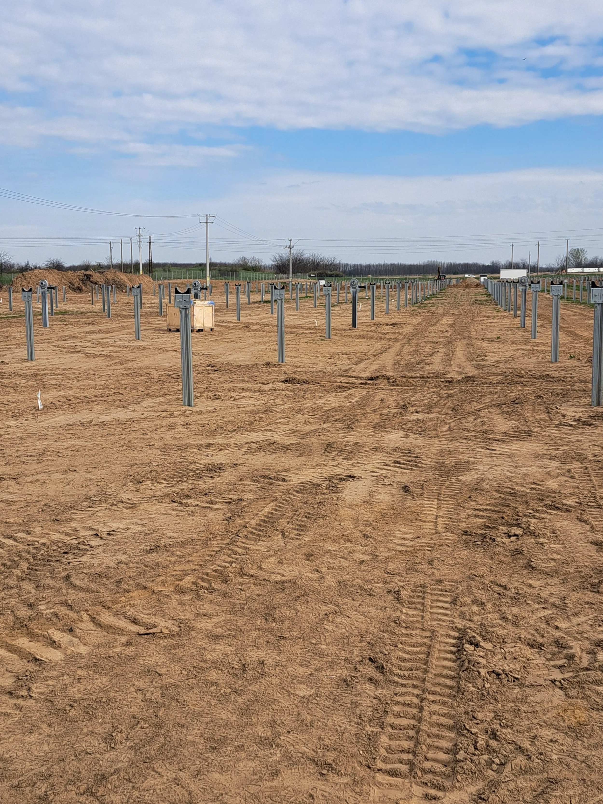 An outdoor scene of a farm under construction, with metal farm equipment or structures in rows in the dirt, and power lines in the background under a partly cloudy sky.
