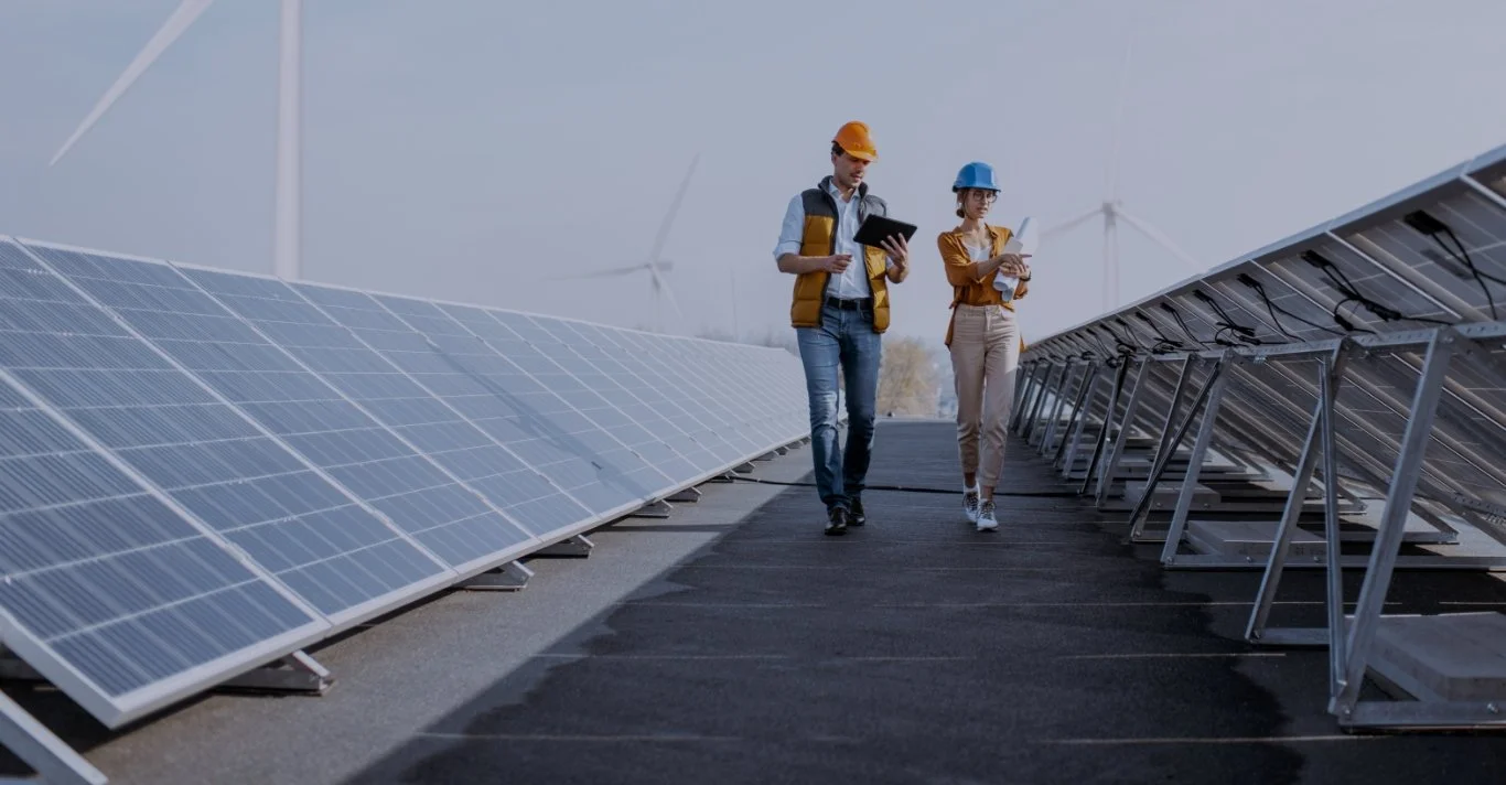 Two engineers inspecting solar panels in a solar farm with wind turbines in the background.