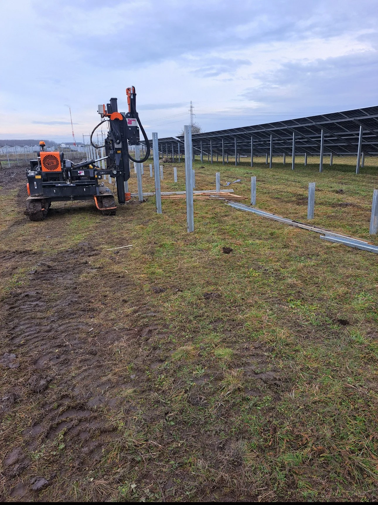 A solar farm with solar panels installed on metal supports. A small construction vehicle, possibly a mini excavator, is in the foreground, working on metal frames for more solar panels. The ground is muddy with patches of grass. The sky is overcast.