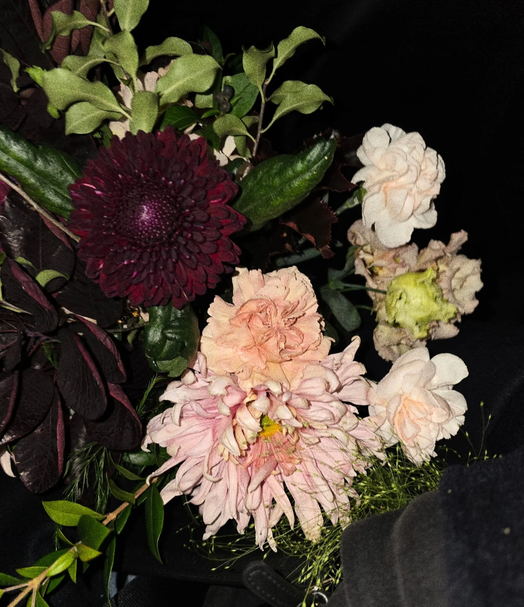 Flower bouquet with burgundy, pink, peach, and white flowers and green foliage on a dark background.