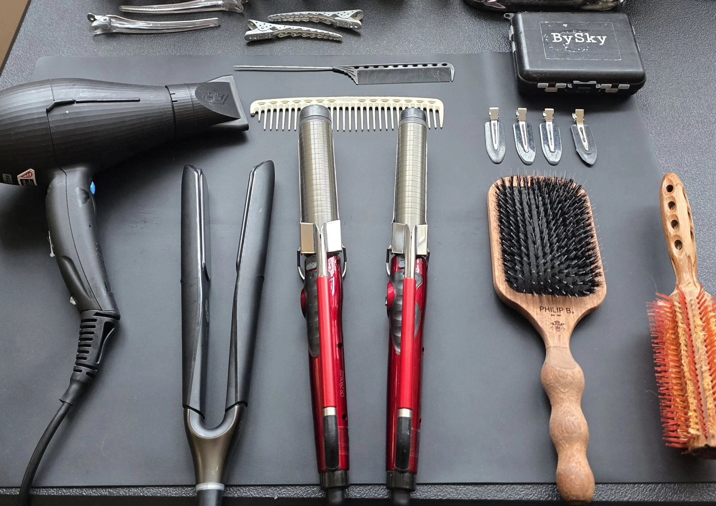 Array of hair styling tools including a hairdryer, hair scissors, thinning shears, combs, hairbrush, and hairbrush cleaner on a black surface.