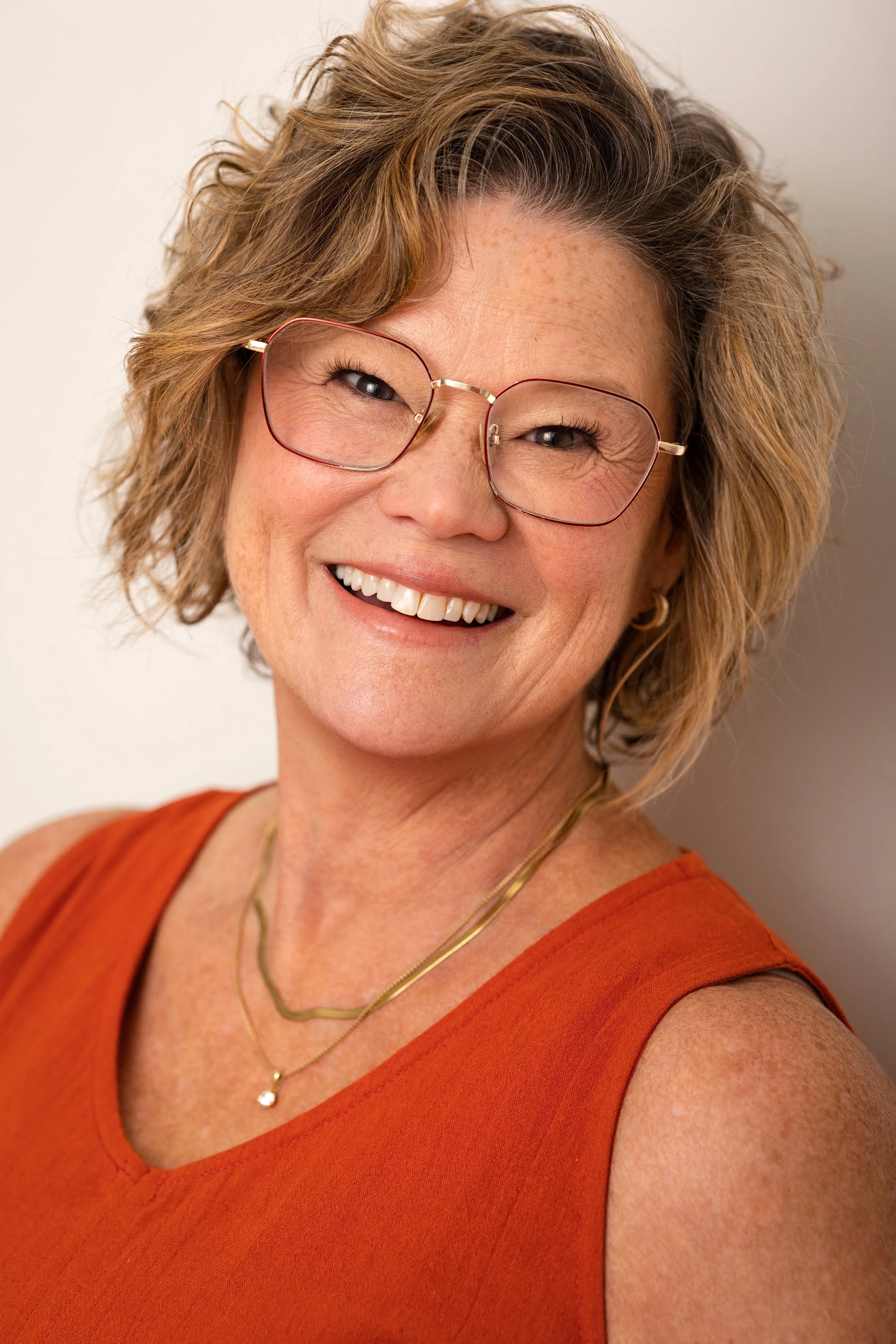 A smiling woman with short, curly light brown hair and glasses, wearing an orange sleeveless top and gold jewelry, standing against a plain white background.
