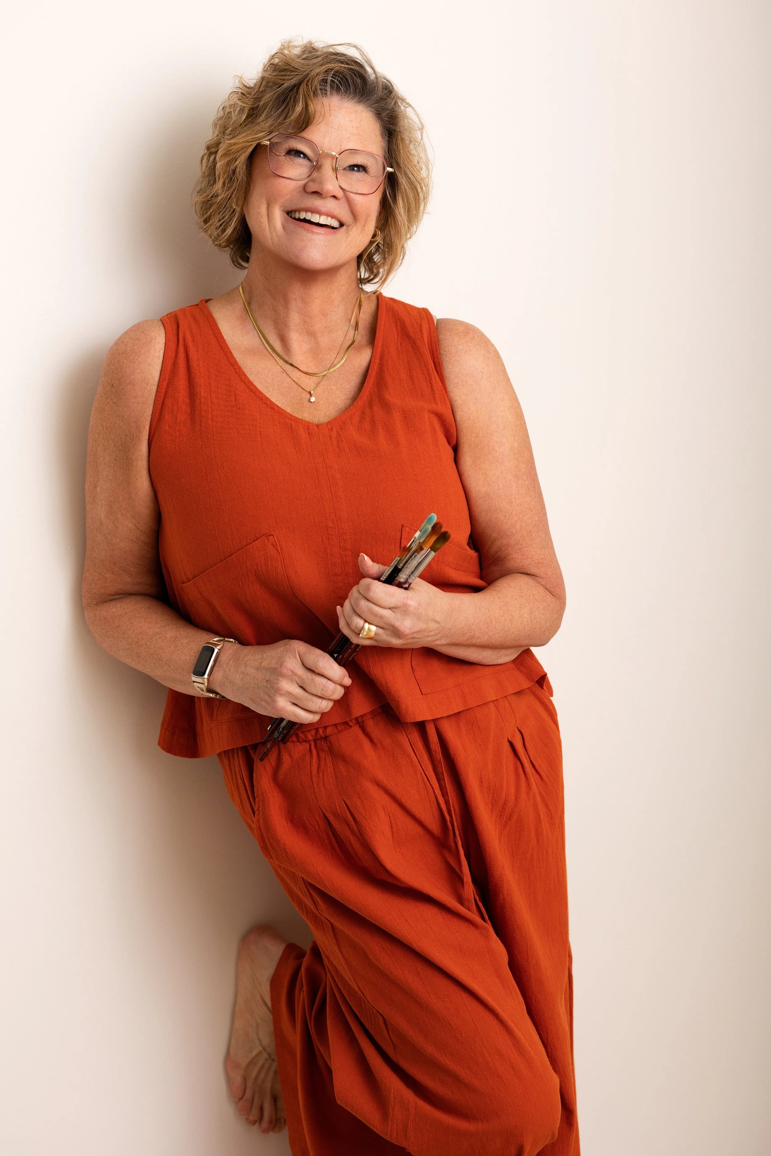 A smiling woman with glasses and short curly hair, wearing an orange sleeveless top and matching pants, holding pens, standing against a plain white wall.