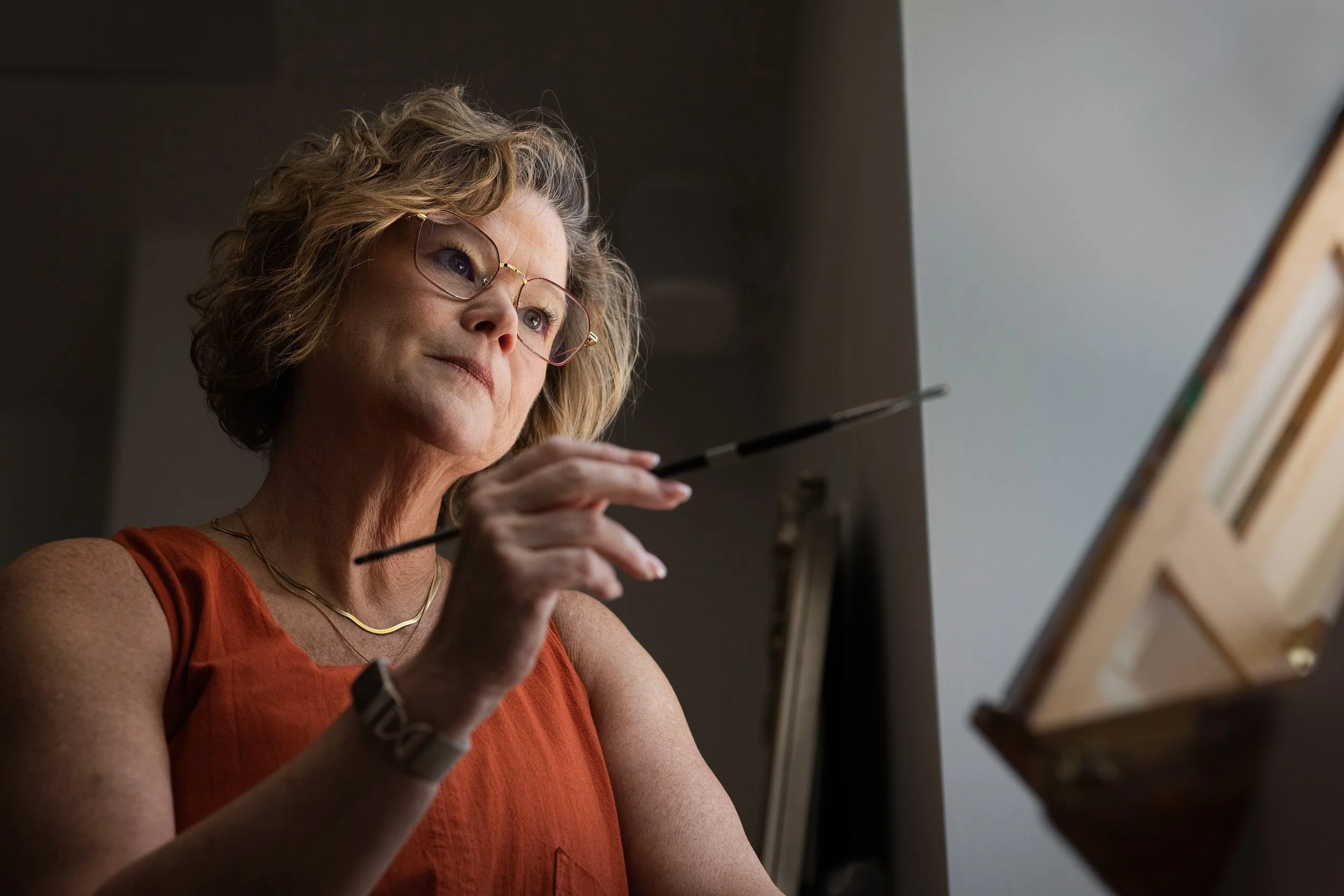 A woman with curly blonde hair wearing glasses and an orange sleeveless top is painting on a canvas in a well-lit room.