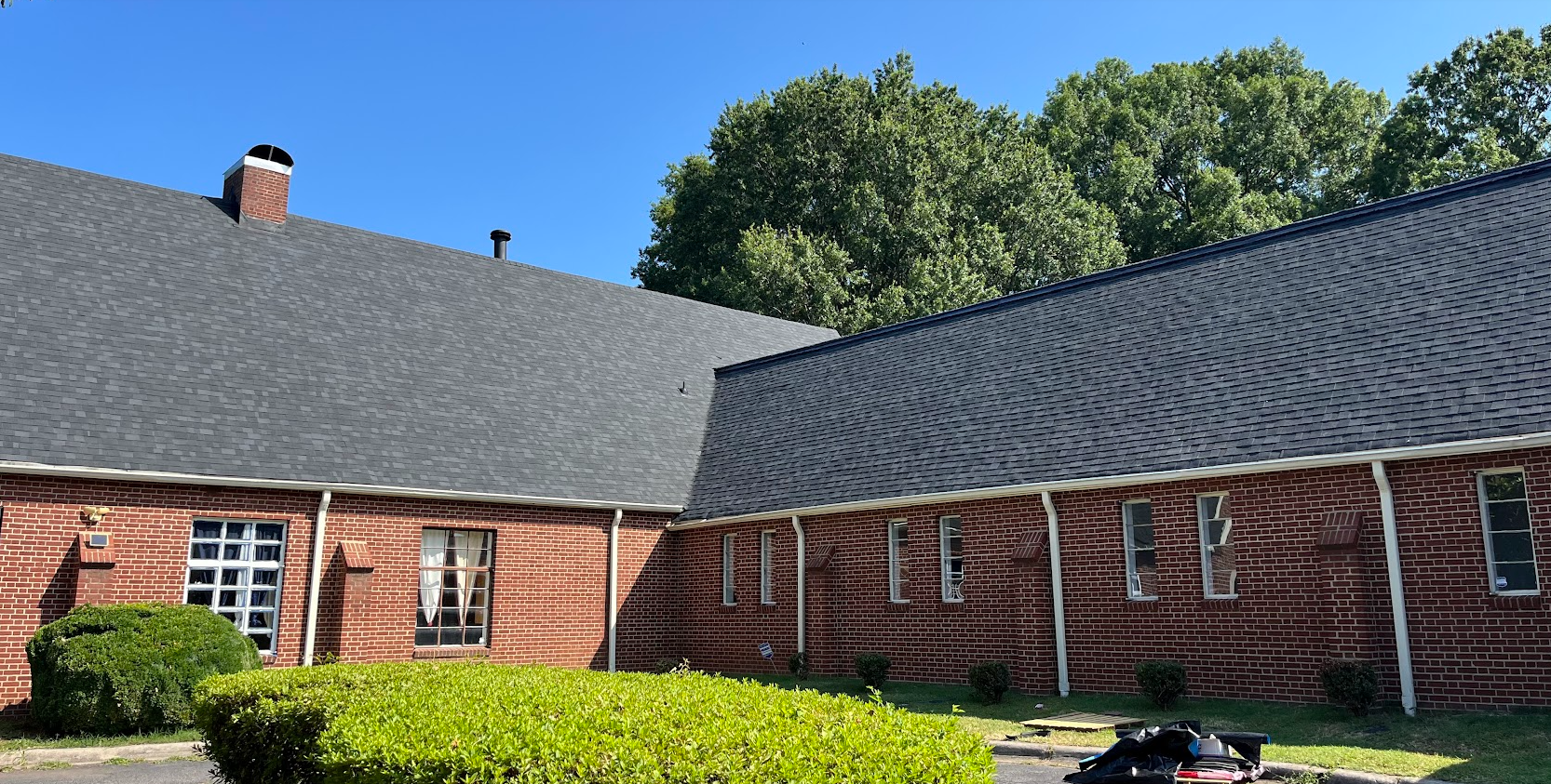 View of a brick building with multiple windows, a sloped shingled roof, and a clear blue sky, with trees in the background and green bushes in the foreground.