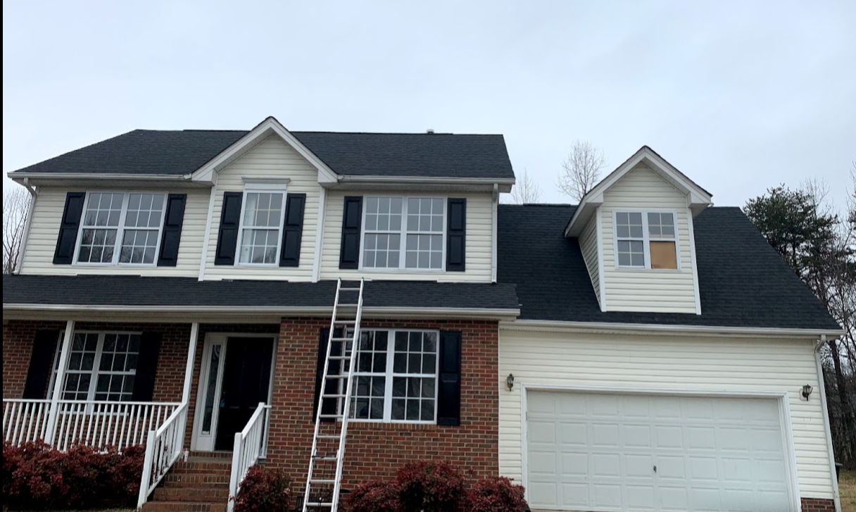 Two-story house with brick and white siding exterior, black shutters, a front porch, and a white garage door. Ladder leaning against the house.