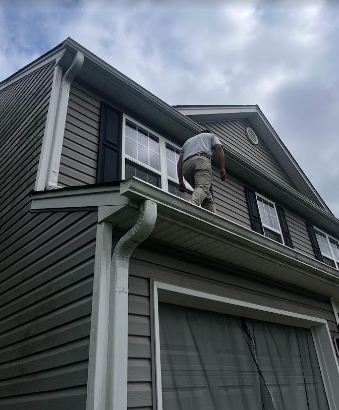 A man working on the roof of a gray, multi-story house with multiple windows and a large window on the ground level, under a cloudy sky.