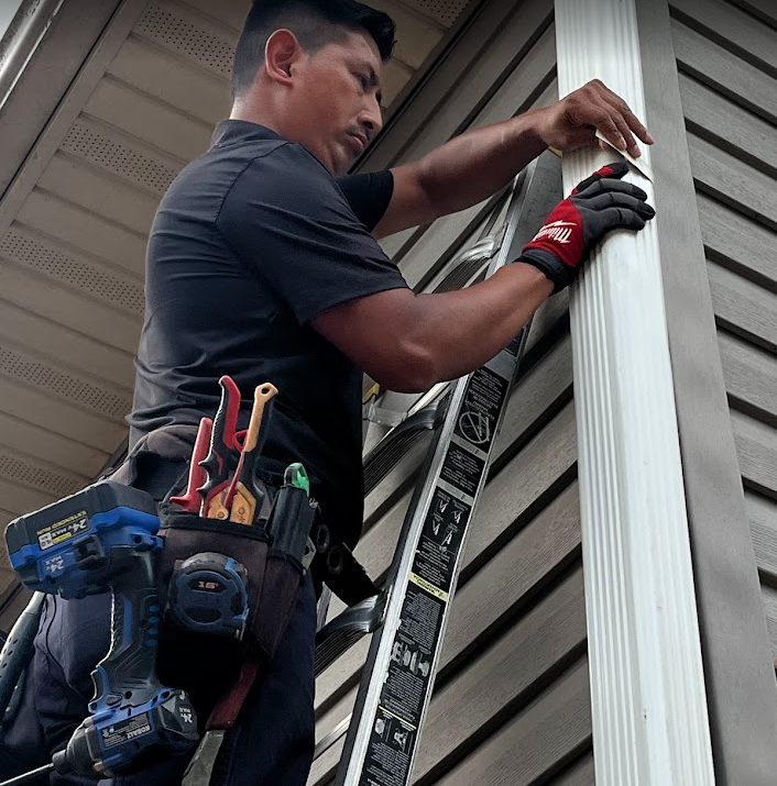 Man working on siding of house, standing on ladder, wearing black shirt, tool belt, and red gloves