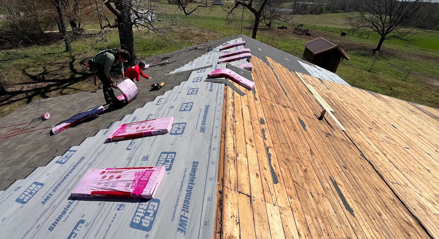 Two workers, one adult and one child, installing new roofing insulation and underlayment on a house roof, with the old wooden shingles partially exposed.