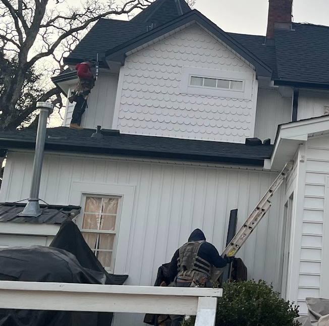 Two workers on ladders repairing or inspecting the exterior of a white house, with one worker on the roof and another on a lower ladder, during daytime.