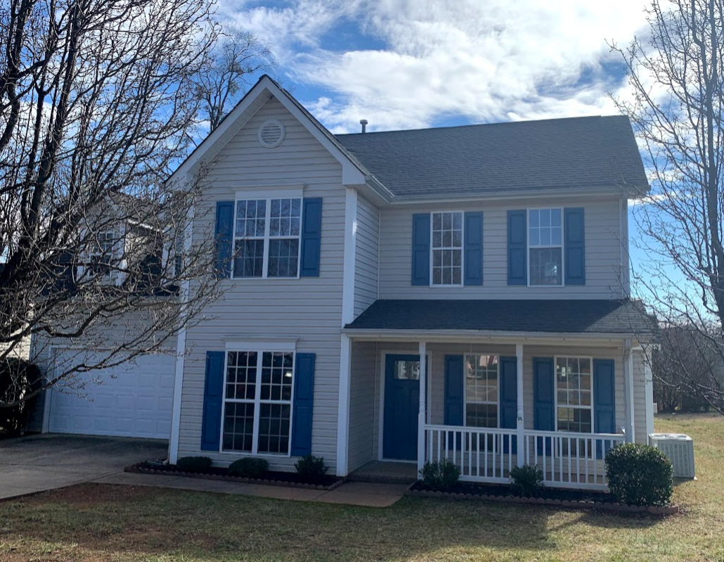 Two-story house with light-colored siding, blue shutters, and a front porch with a small white railing, surrounded by a lawn with some shrubs, and leafless trees in the front yard.