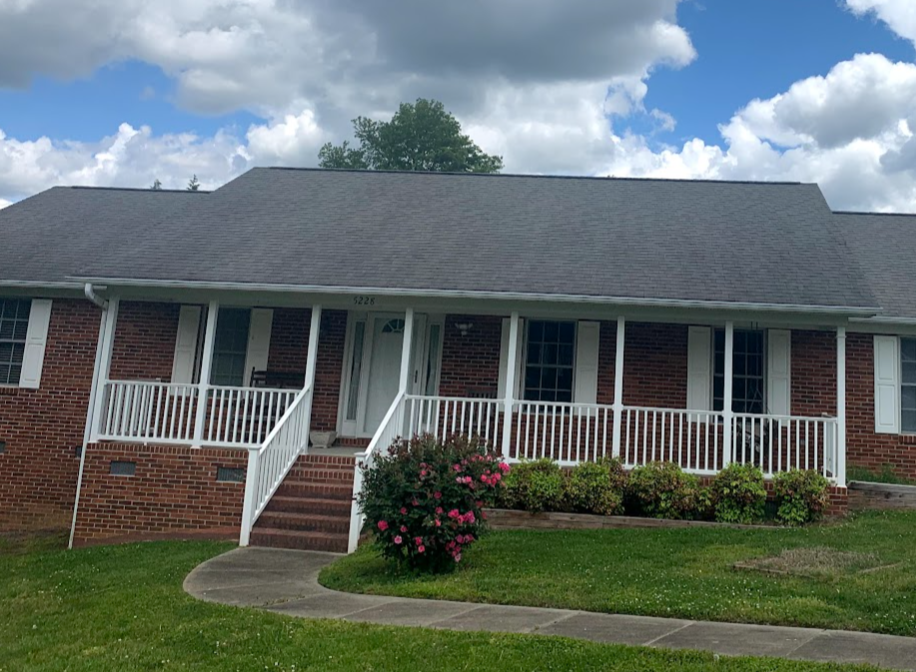 Brick house with white porch railing, steps, and multiple windows, surrounded by grass, pink flowering bush, and cloudy sky.