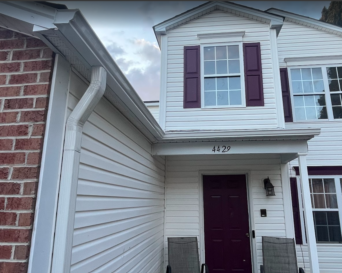 Front entrance of a two-story house with white siding, purple shutters, and a dark purple door. Two chairs are on the porch, and the house number 4429 is above the door. A brick wall and gutter are visible on the left side.