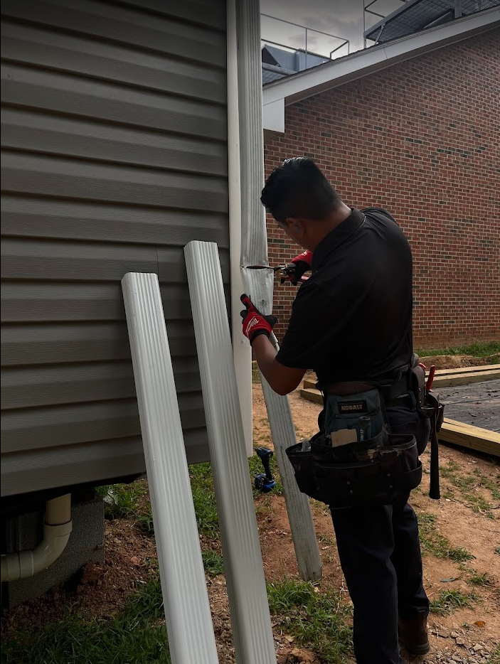 A person installing siding on a house exterior using a tool, with several metal support poles nearby.