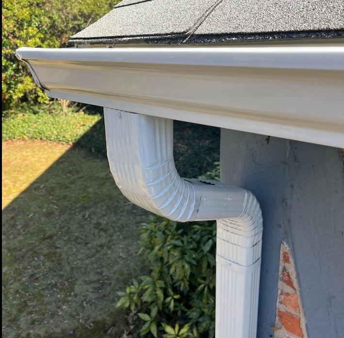 Close-up of white gutter and downspout attached to the side of a house with gray exterior siding and a brick foundation, with a garden and grass in the background.