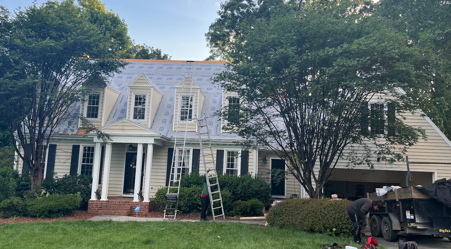 A house under renovation with new roofing being installed. Ladders are set up on the front yard, and workers are working near a trailer in the driveway. The house is surrounded by trees and bushes, with a well-maintained lawn in the foreground.