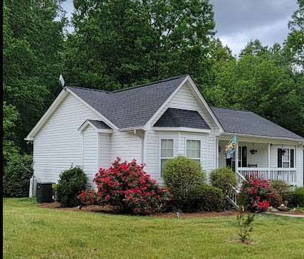 A white house with a gray roof, surrounded by green trees and bushes with pink flowers, on a grassy lawn.