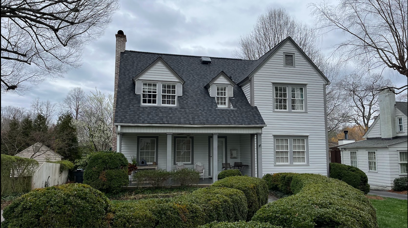 A two-story white house with a dark gray roof, front porch, and multiple windows, surrounded by trimmed bushes and trees, under a cloudy sky.