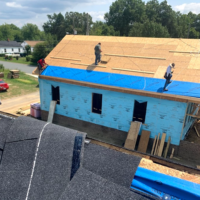 Construction workers installing a new roof on a house during daytime, with some workers on the roof and blue roofing underlayment visible.