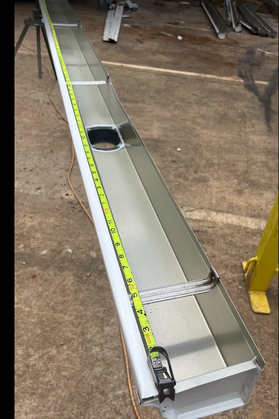 Aluminum metal structure with a measuring tape stretched along its length, placed on a concrete floor in a workshop or construction area.