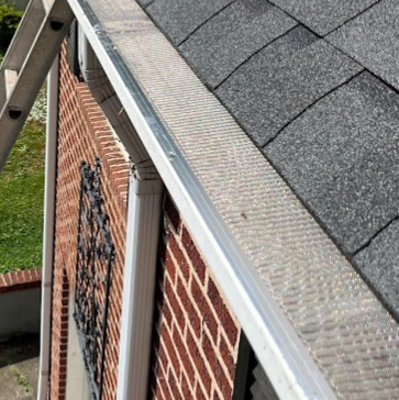 Close-up of a house's roof edge with roofing shingles, gutter, and brick wall in the background.