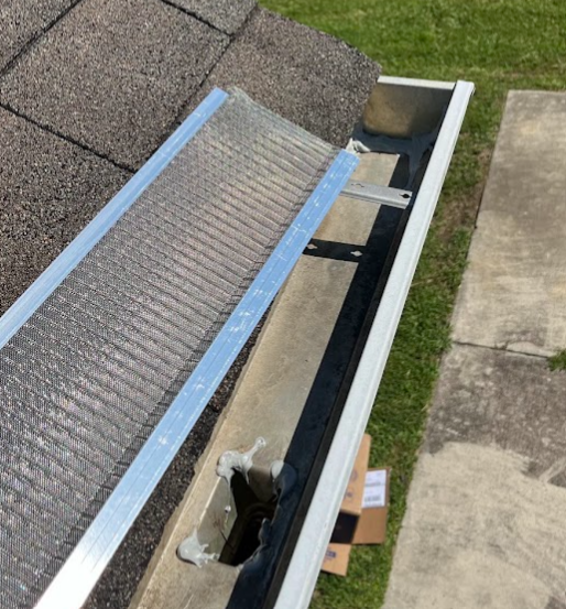 Close-up of a gutters assembly attached above a concrete sidewalk with a section of mesh screen, a ruler, and a cardboard box nearby.