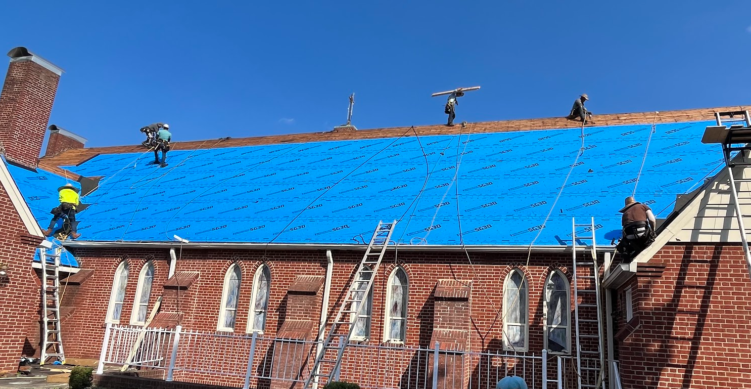 Construction workers installing blue roofing underlayment on a brick church roof under a clear blue sky, with ladders and safety equipment visible.