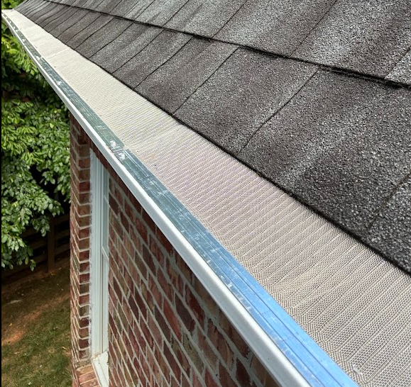 Close-up of a house roof's edge showing shingles and a metal gutter with leaf guard, attached to a brick wall.