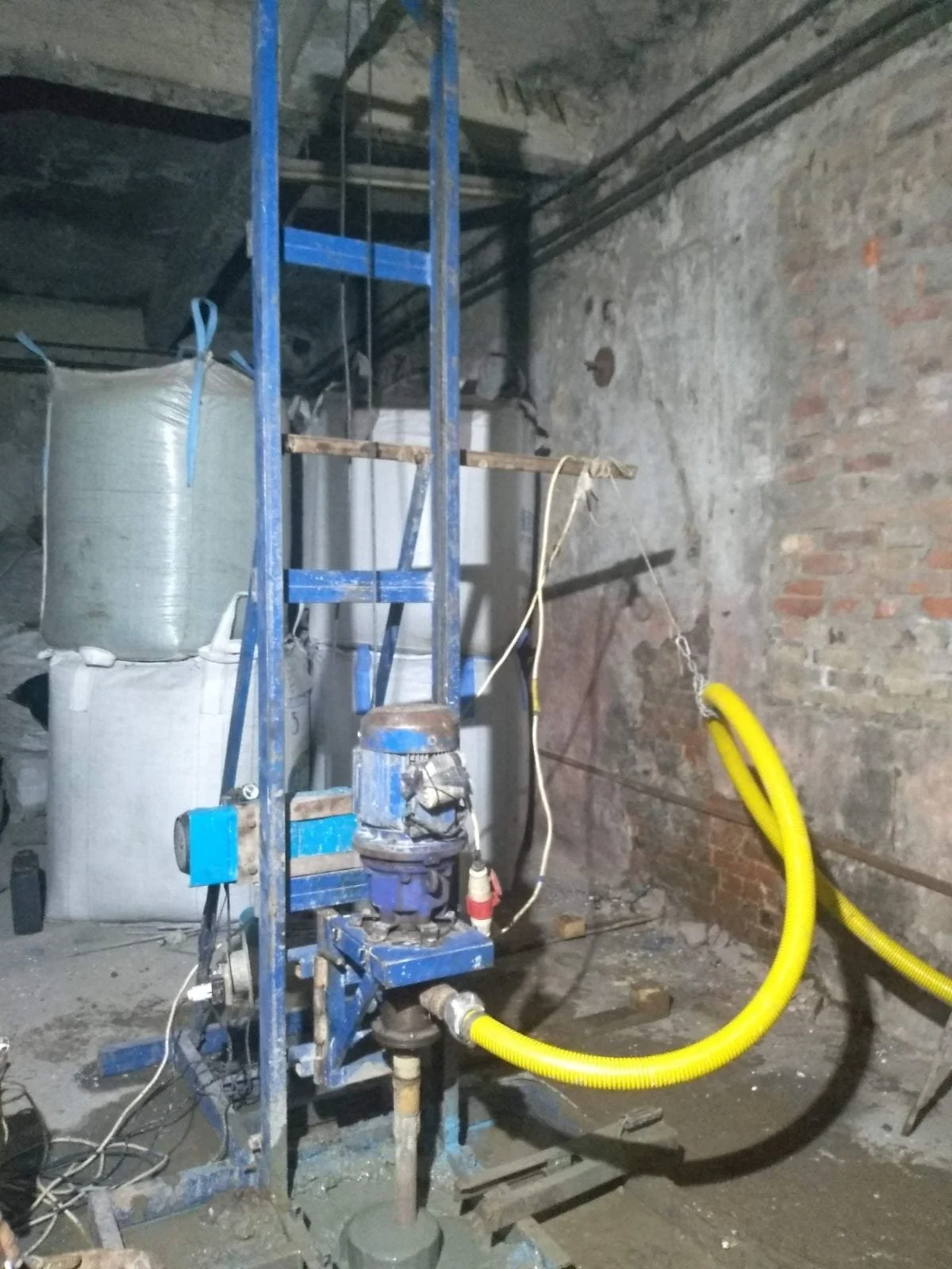 Construction site in an unfinished building with exposed brick walls and construction equipment, including a blue scaffolding, yellow and white flexible hoses, and large bags stored on the left side.