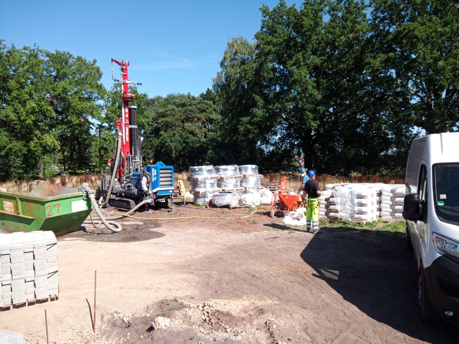 Construction site with workers in safety gear, pallets of materials, and a drilling machine