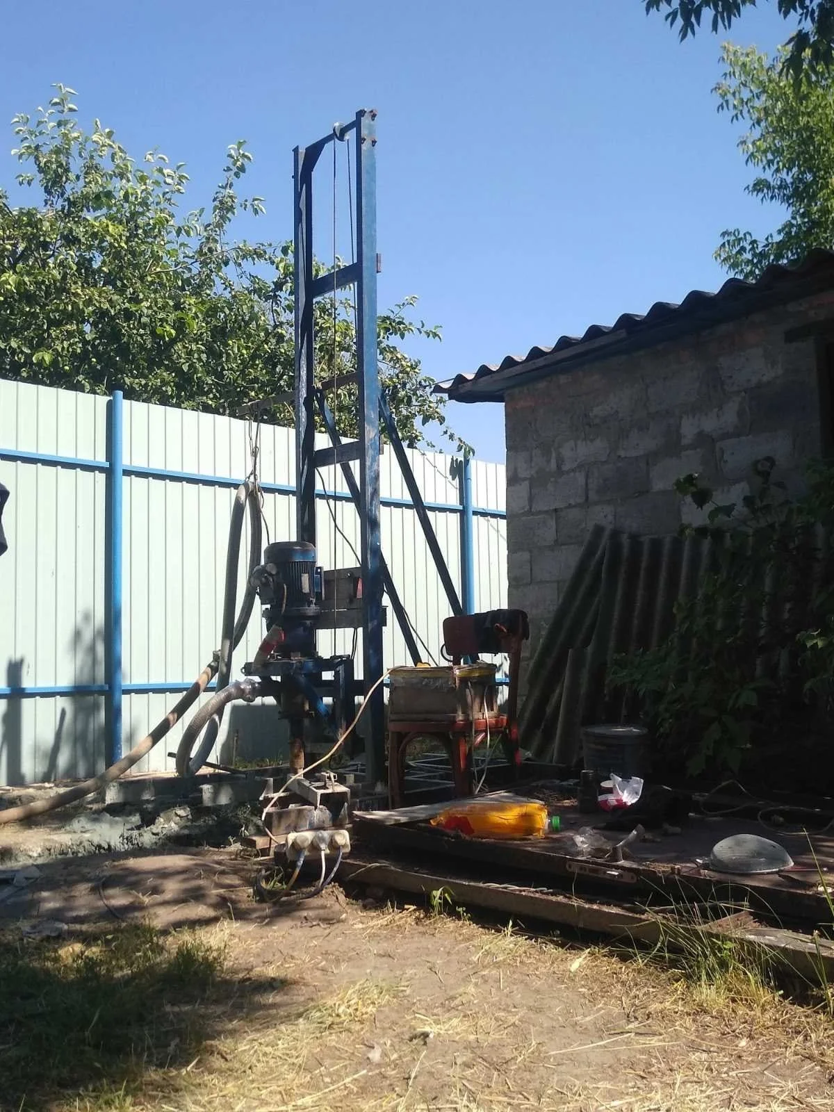 A construction site with a tall blue drilling machine, a concrete block building, metal pipes stacked against a wall, and various tools and objects on the ground, under a clear blue sky.
