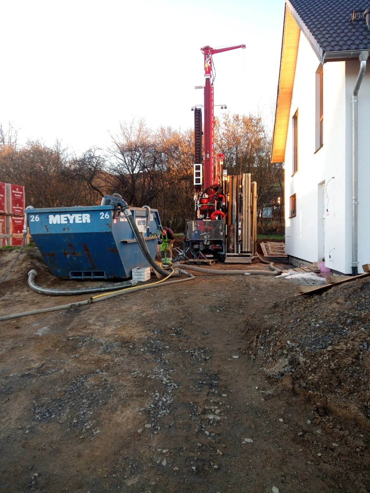 Construction site near a white house with a dark roof, showing construction equipment including a blue container labeled 'MEYER', a worker in safety gear, and a tall red crane with various pipes and machinery.