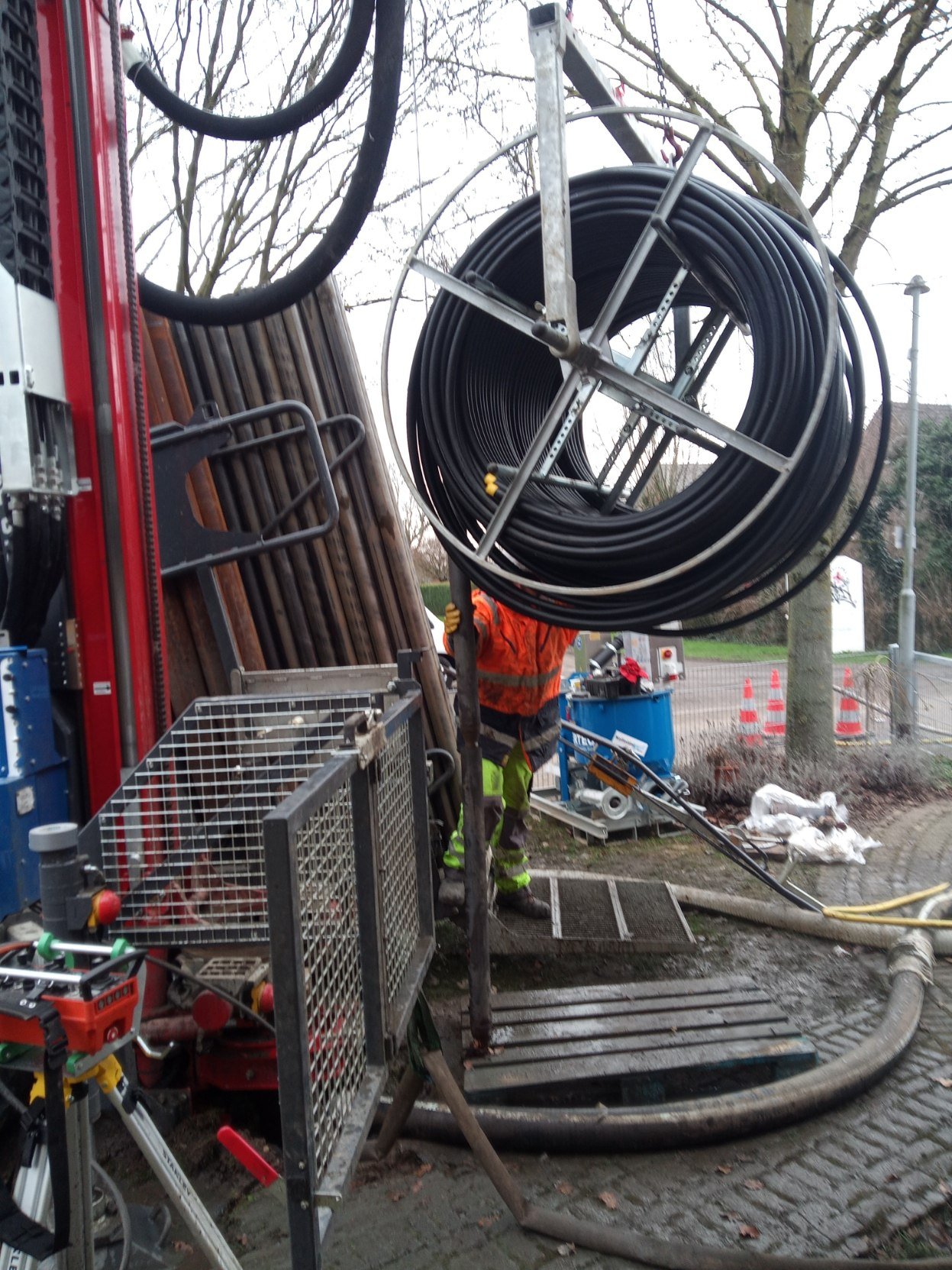 Construction workers installing large underground pipes with a crane and equipment, set outdoors near trees and a sidewalk.