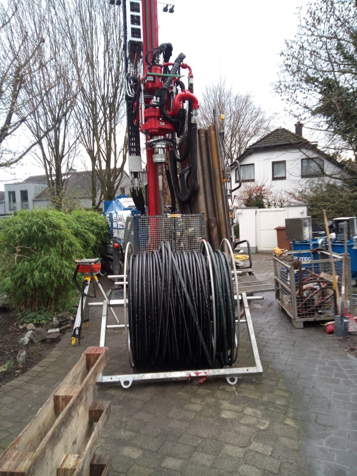 A large red hydraulic drilling machine equipped with hoses and pipes, with coiled black cables in front, placed on a paved driveway in a residential area with houses, trees, and gardening equipment nearby.