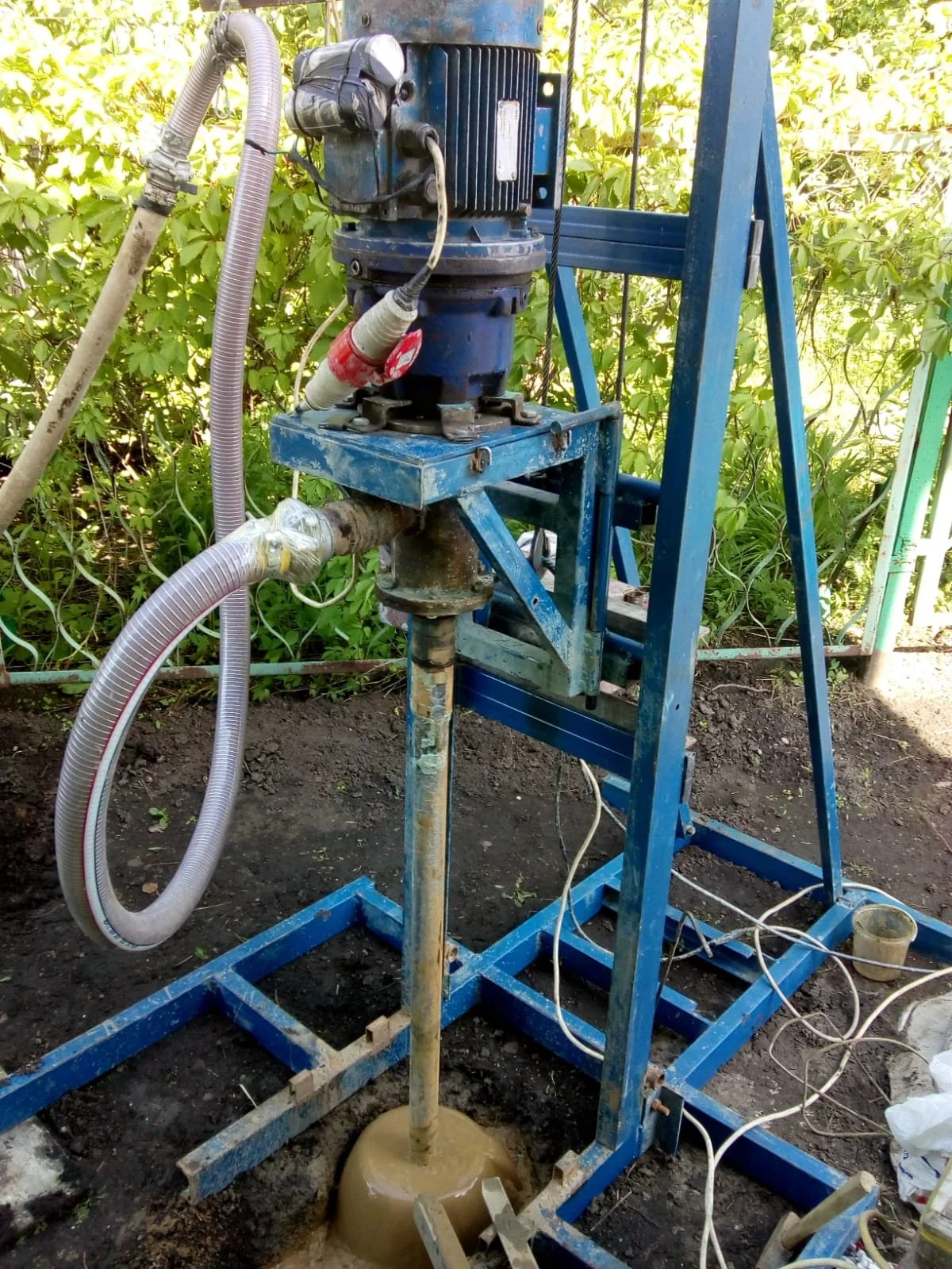 A blue construction or industrial machine with hoses connected, standing outdoors on dirt soil, surrounded by green foliage, with electrical wires and a small container nearby.